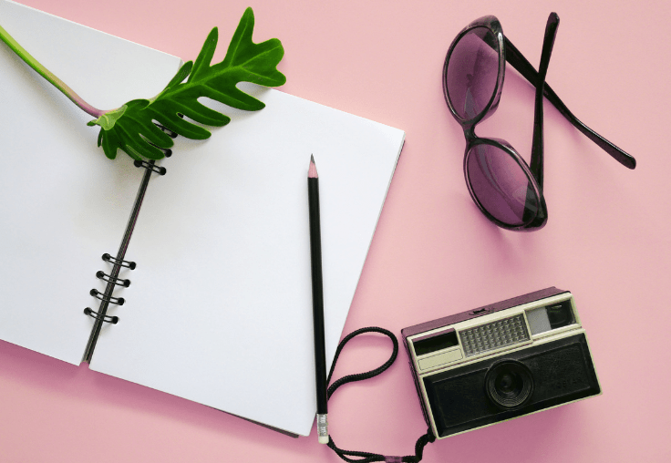 vignette of a notebook with a leaf and a pencil on it, a pair of sunglasses in the upper right and a camera on the lower right