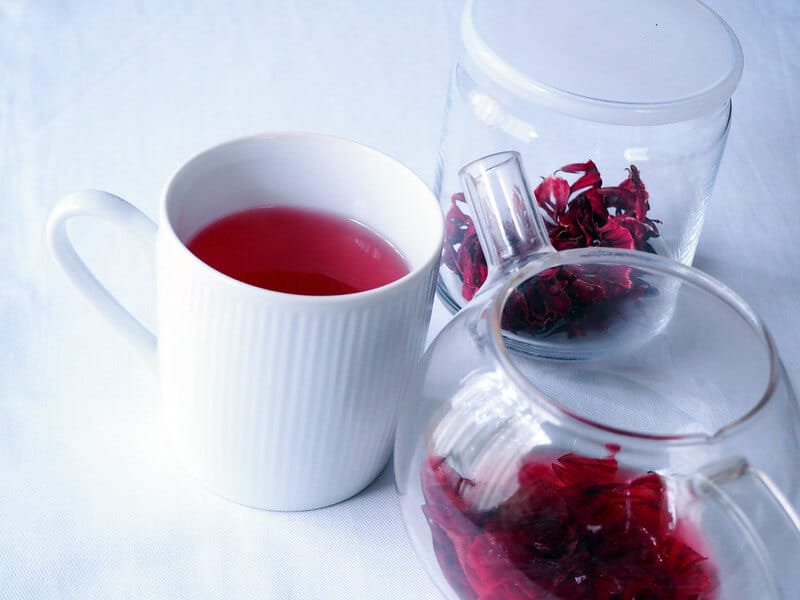White coffee mug with roselle tea in it, a crystal teapot with tea in it and a crystal bowl with roselle calyces